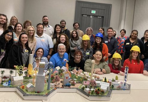 Group photo of participants wearing hard hats, proudly posing behind their completed miniature city during a City Build team building event.
