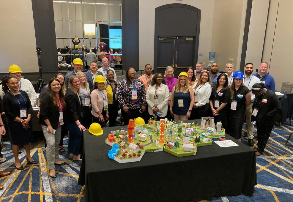 Team members in hard hats pose together around a completed City Build project, showcasing a colorful miniature city model with roads, buildings, and greenery.