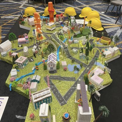 A completed City Build team building project featuring a vibrant miniature city with buildings, roads, trees, and glowing lights, surrounded by yellow hard hats on display. thumbnail