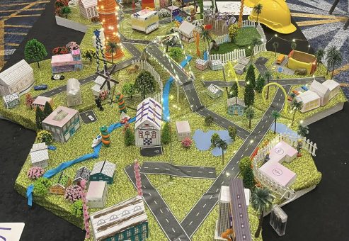 A completed City Build team building project featuring a vibrant miniature city with buildings, roads, trees, and glowing lights, surrounded by yellow hard hats on display.