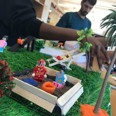 A close-up of a miniature playground scene at a City Build team building event, featuring toy figurines, a sandbox, palm trees, and vibrant greenery, with participants working in the background. thumbnail