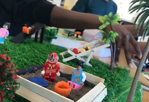 A close-up of a miniature playground scene at a City Build team building event, featuring toy figurines, a sandbox, palm trees, and vibrant greenery, with participants working in the background.