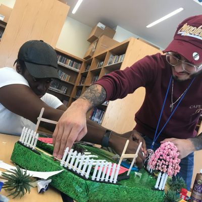 Two participants collaborate to build a detailed section of a miniature city during a City Build team building activity, adding fences and trees to the green landscape. thumbnail