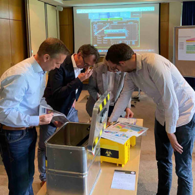 Group of participants analyzing documents and tools around a biohazard-themed box as part of the 'Atmosphere ' team building activity. thumbnail