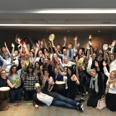 A large group of participants celebrating together after a successful BeatsWork team building session, holding percussion instruments and posing with enthusiasm indoors. thumbnail