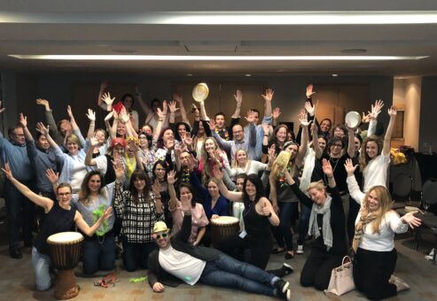 A large group of participants celebrating together after a successful BeatsWork team building session, holding percussion instruments and posing with enthusiasm indoors.
