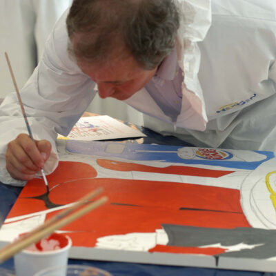A participant wearing a white protective suit carefully paints a section of a mural during The Big Picture team building event. The individual focuses on adding details to a vibrant red, blue, and white canvas, contributing to the collaborative masterpiece. thumbnail