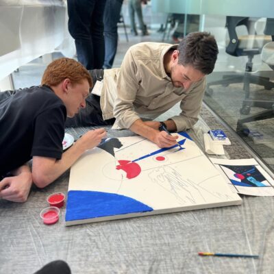 Two participants are lying on the floor, working together to paint a section of the canvas during The Big Picture team building event. They focus intently on adding details and colors, collaborating on their contribution to the larger mural. thumbnail