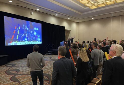 Participants in a Boom Time team building event are actively following a colorful rhythm-based music display projected on a large screen. Holding Boomwhackers, they collaborate to play the correct notes, guided by the display, in a fun and engaging team activity.