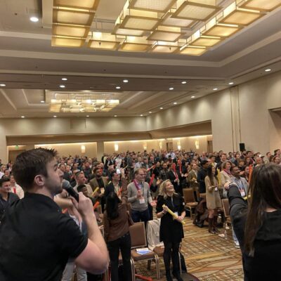 A large group participates in the Boom Time team building activity, guided by energetic facilitators. The crowd enthusiastically follows along with musical cues, using colorful Boomwhackers to create rhythmic sounds together. thumbnail
