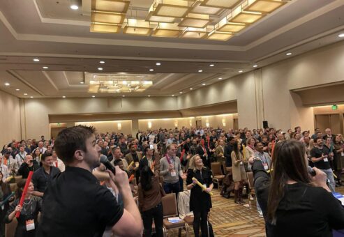 A large group participates in the Boom Time team building activity, guided by energetic facilitators. The crowd enthusiastically follows along with musical cues, using colorful Boomwhackers to create rhythmic sounds together.