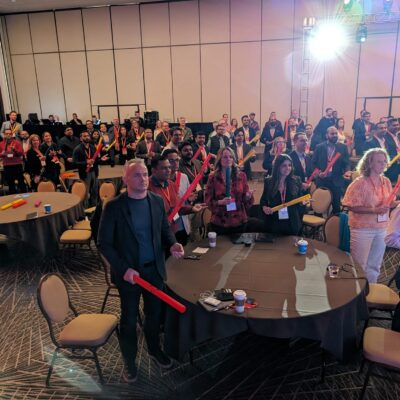 Conference attendees stand at their tables holding colorful Boomwhackers, engaged in a fun, interactive Boom Time team building session, focused on rhythm and collaboration. thumbnail
