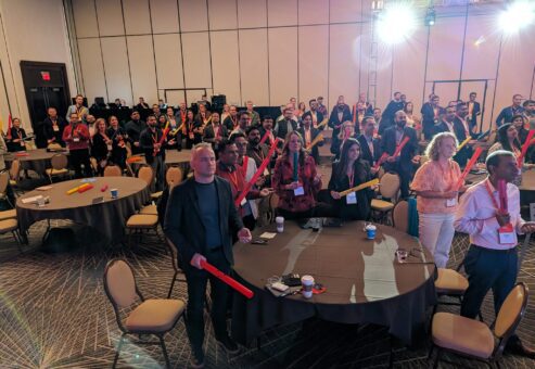 Conference attendees stand at their tables holding colorful Boomwhackers, engaged in a fun, interactive Boom Time team building session, focused on rhythm and collaboration.