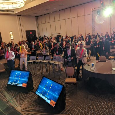 Conference participants engage in a high-energy Boom Time team building session, using colorful Boomwhackers to follow musical patterns displayed on large screens. The event promotes collaboration, rhythm, and excitement in a lively setting. thumbnail