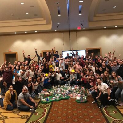 Large group celebrating their completed City Build project during a team building event, with a colorful miniature city displayed in the center of the room. thumbnail