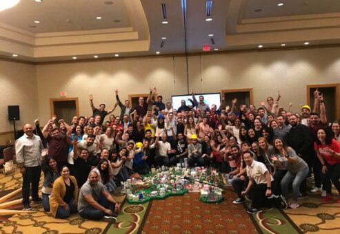 Large group celebrating their completed City Build project during a team building event, with a colorful miniature city displayed in the center of the room.