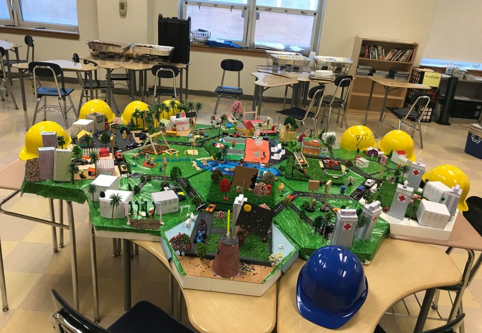 Colorful miniature city model displayed on tables, surrounded by yellow and blue hard hats, featuring buildings, roads, and greenery for a City Build team building activity.
