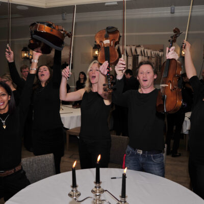 Excited participants raise their violins triumphantly after performing together during the Crescendo team building event. thumbnail