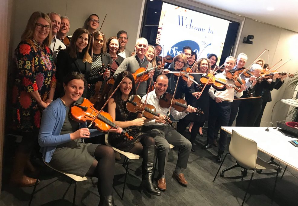 Group of participants playing violins together in a meeting room during the Crescendo team building event, smiling and enjoying the collaborative musical activity.