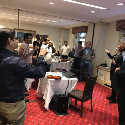 Participants in a Crescendo team building event stand in a circle, holding violins and bows, as an instructor leads them through a collaborative music lesson in a conference room. thumbnail