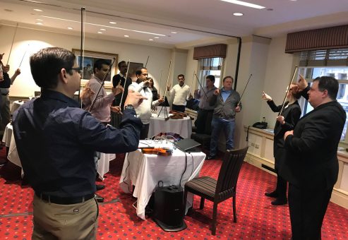 Participants in a Crescendo team building event stand in a circle, holding violins and bows, as an instructor leads them through a collaborative music lesson in a conference room.