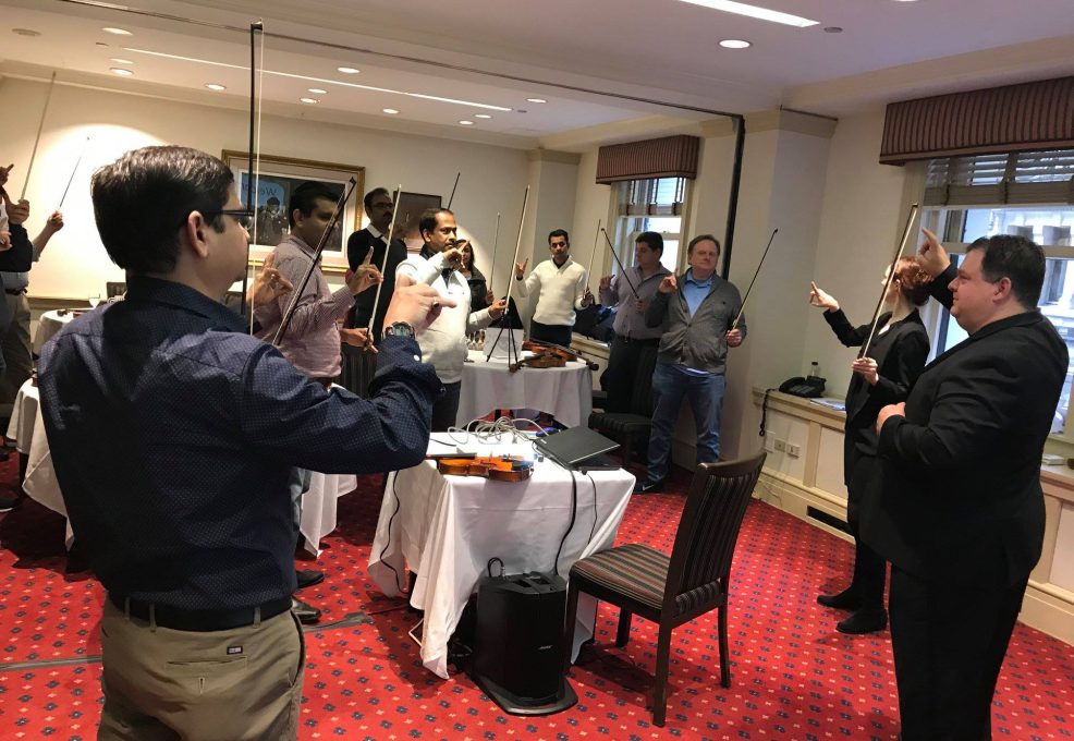 Participants in a Crescendo team building event stand in a circle, holding violins and bows, as an instructor leads them through a collaborative music lesson in a conference room.