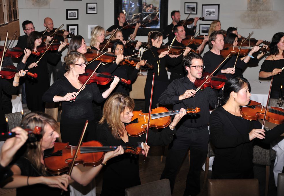 Participants in black clothing perform together as a string orchestra during a Crescendo team building event, holding violins and violas with focused expressions.