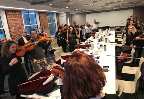 Participants in a Crescendo team building event practicing their violins together in a bright, modern conference room, guided by instructors.