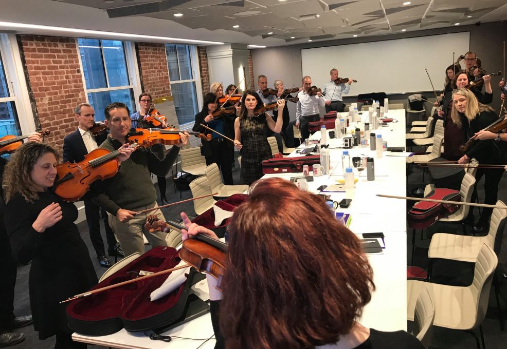 Participants in a Crescendo team building event practicing their violins together in a bright, modern conference room, guided by instructors.