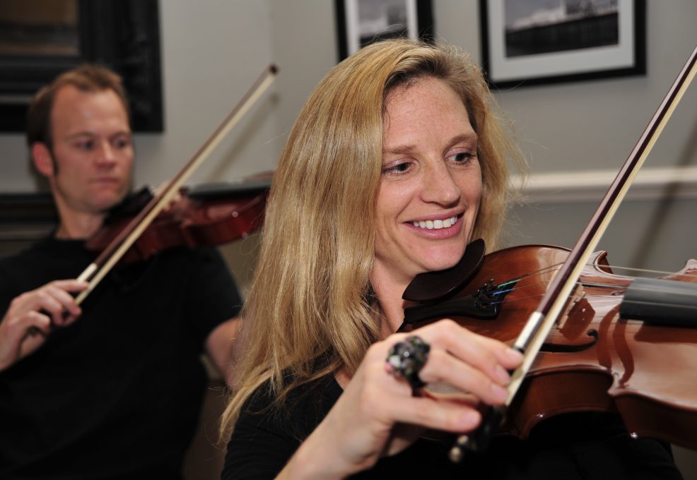 Smiling participants learning to play violins together during the Crescendo team building event, highlighting teamwork and musical collaboration.