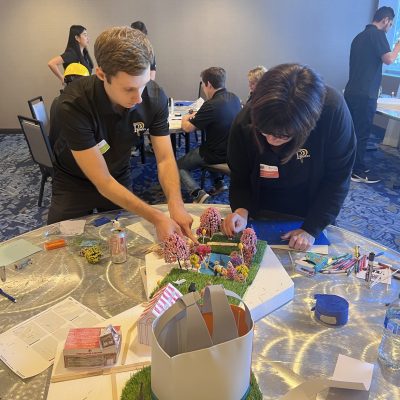 Two participants at a City Build team building event focus on constructing a detailed miniature city model using materials like trees, buildings, and flags. thumbnail