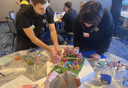 Two participants at a City Build team building event focus on constructing a detailed miniature city model using materials like trees, buildings, and flags.