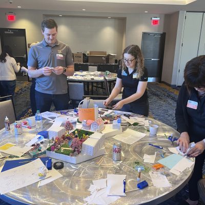 Team members at a City Build team building event collaborate to design and assemble a creative miniature city model using various materials on a round table. thumbnail