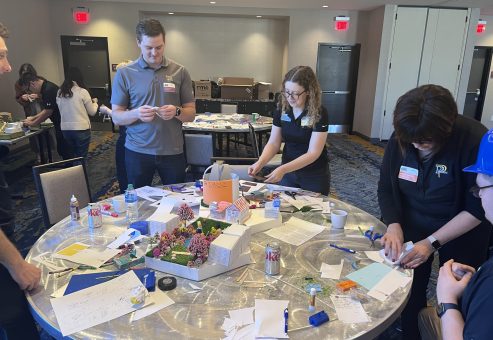 Team members at a City Build team building event collaborate to design and assemble a creative miniature city model using various materials on a round table.