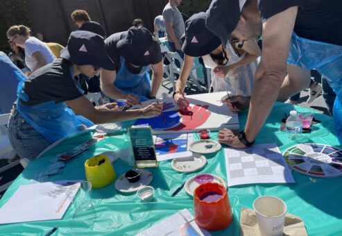 A group of participants in matching hats and aprons collaborate on painting a vibrant section of a mural during The Big Picture team building event. They work together, adding colors to the canvas under a sunny outdoor setting, with painting supplies and color wheels scattered on the table.