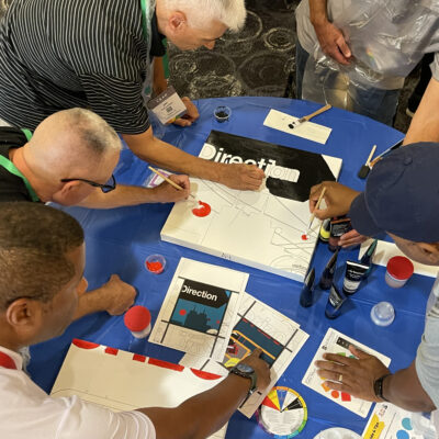 A group of participants works together painting sections of a mural during The Big Picture team building event, focusing on precision and collaboration to create a unified artwork. thumbnail