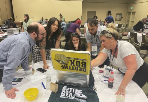 Team members gather around a challenge box during the Beat the Box team building event, strategizing together to solve escape room-style puzzles and unlock their way to victory.