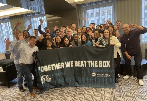 A large group of excited participants celebrate their victory after successfully completing the Beat The Box team building challenge, proudly displaying their teamwork banner.