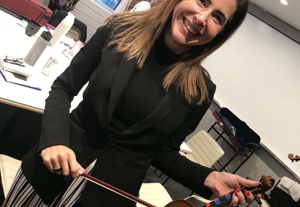 Smiling participant holds a violin and bow during the Crescendo team building event, standing in a meeting room with tables and chairs.