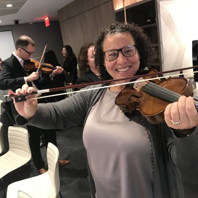 Smiling participant holds a violin and bow proudly during the Crescendo team building event, surrounded by teammates learning to play instruments. thumbnail