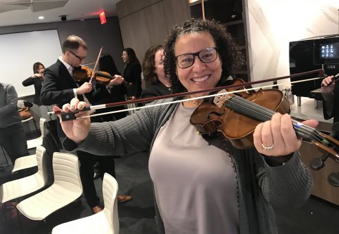 Smiling participant holds a violin and bow proudly during the Crescendo team building event, surrounded by teammates learning to play instruments.