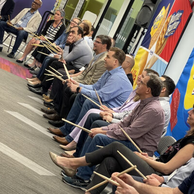 A group of participants seated in a row, holding drumsticks and preparing for a rhythm-based activity during a BeatsWork team building event. Everyone is focused and ready to engage in the upcoming musical challenge. thumbnail