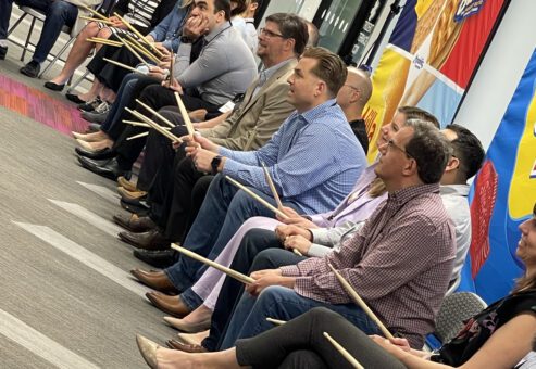 A group of participants seated in a row, holding drumsticks and preparing for a rhythm-based activity during a BeatsWork team building event. Everyone is focused and ready to engage in the upcoming musical challenge.