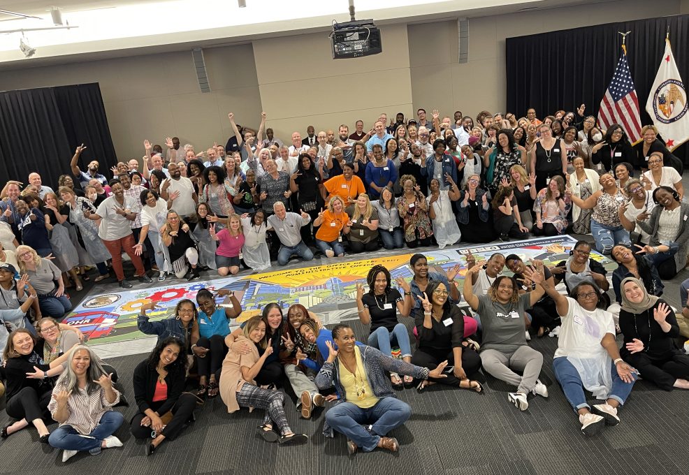A large group of participants celebrates completing a collaborative mural during The Big Picture team building event, showcasing teamwork and creative achievement.
