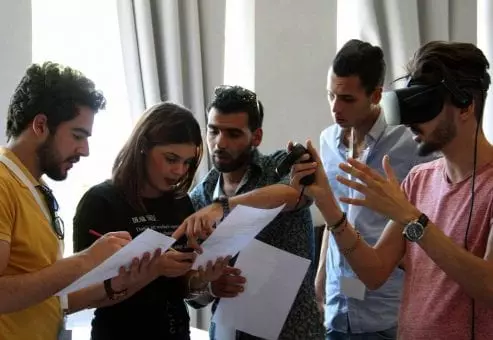 A group of participants collaborates on a task, with one member wearing a VR headset and explaining the virtual challenge to the rest of the team during The Infinite Loop team building event.