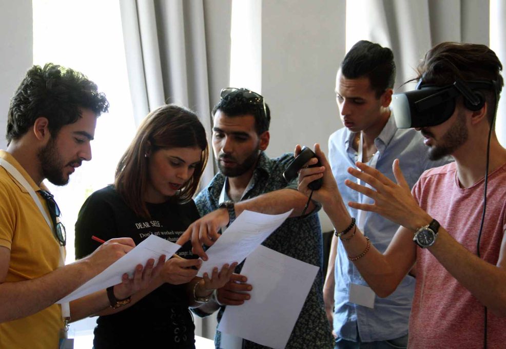 A group of participants collaborates on a task, with one member wearing a VR headset and explaining the virtual challenge to the rest of the team during The Infinite Loop team building event.