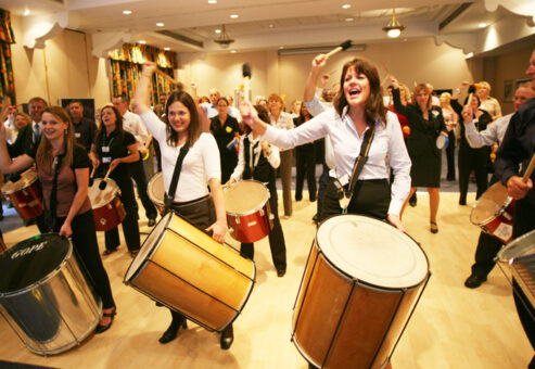 A group of energetic participants playing large percussion drums during a lively indoor BeatsWork team building session. They are smiling and engaged, enjoying the rhythm and teamwork experience.