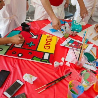 Participants work together on a colorful mural panel during The Big Picture team building event. The table is filled with paint supplies, and one of the participants carefully paints a section featuring bold shapes and colors. thumbnail