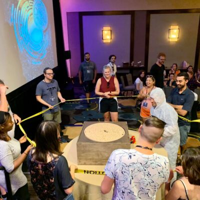 Group of participants gathers around a large maze puzzle table, focusing on solving the challenge together during the 'Escape The Maze' team building event, surrounded by caution tape for an immersive experience.