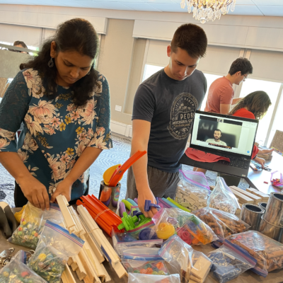Participants sorting through materials and building supplies during the Domino Effect team building event, with a remote team member visible on a tablet. thumbnail